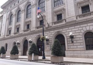 11th circuit - NARSOL Stone government building with arched windows, a street lamp bearing an American flag, and large potted evergreens along the sidewalk.