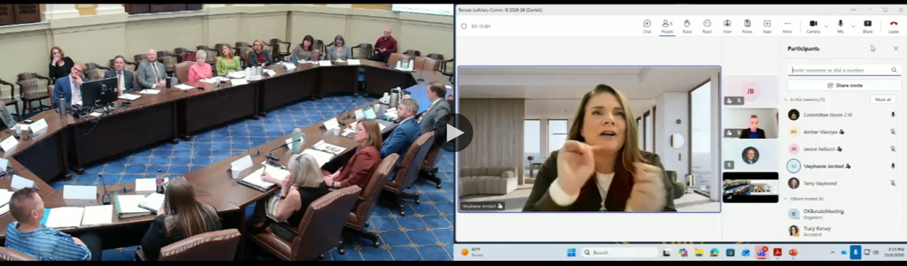 Large conference room with attendees around a curved table; a video call window shows a woman presenting remotely.