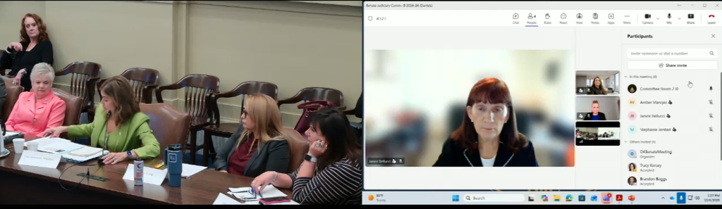 Hybrid meeting: in-person group of women at a long table on the left and a video conference window showing a woman speaker on the right.