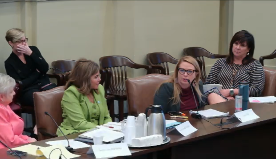 Five women sit around a conference table with microphones, papers, and coffee mugs, engaged in discussion during a meeting.",