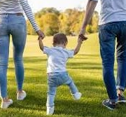 parent-child - NARSOL Toddler between two adults holding hands, walking on a sunlit grassy park.